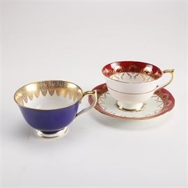 Aynsley Bone China Serviceware Collection: A collection of Aynsley bone china serviceware. This three-piece grouping includes one footed cup in the 1713 pattern featuring a cobalt blue exterior, a pink rose design to the interior bowl, and a white interior with a gold tone filigree border to the interior rim. Also included is one footed cup and saucer in the 2146 red scalloped pattern featuring a maroon and white palatte, a pink rose design to the interior bowl, and a maroon interior edge with a gold tone filigree border to the interior rim. Each piece is marked to the underside reading “Aynsley England Bone China”.