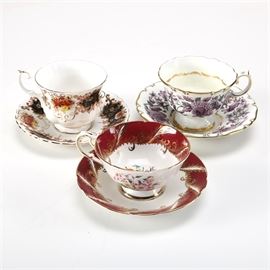 Bone China Tea Cups and Saucers: A trio of fine bone china tea cups and saucers. Offered is a Paragon teacup and saucer with maroon, gold-tone, and floral accents, a Royal Albert Heritage teacup and saucer with a black, gold-tone, maroon, and yellow floral design, and a Grosvenor bone china teacup and saucer set with a green, gold-tone, and violet floral design. Each piece is marked to the bottom with its maker.