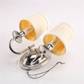 Candelabrum Wall Sconce: A candelabrum wall sconce. The chrome light fixture has an oval backplate and two curved arms with single socket receptacles. The wall sconce includes two pleated linen shades.