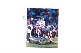 John Elway Signed Photograph: A John Elway signed photograph. It depicts the quarterback setting up a play on field. The color image is hand signed by Elway in in blue. It comes with a protective plastic sleeve.