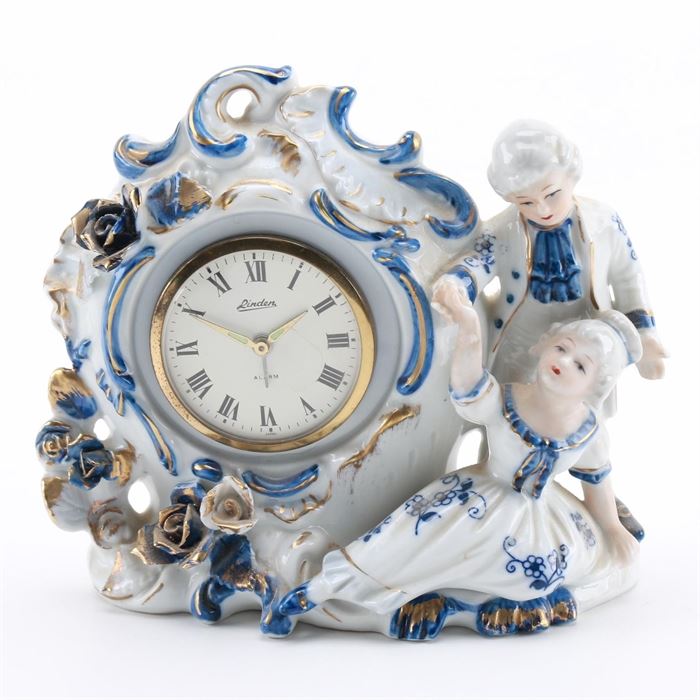 Linden Porcelain Figural Desk Clock: A vintage Linden porcelain figural alarm clock. This clock features white porcelain with floral designs around the clock face, a male and female figure sit at the side and it is finished with blue and gold tone accents. The clock has a wind-up key and is marked “Linden” and “Alarm” on the face and “Japan” on the back, with “Genuine Porcelain China” to the underside.