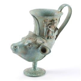 Replica Etruscan Drinking Vessel by Riproduzione Archeologica: A Riproduzione Archeologica replica of an Etruscan drinking vessel. The ornate cup features a cow head with Etruscan god images to each side on a pedestal base. The piece is marked to the underside “Riproduzione Archeologica, Made in Italy.”
