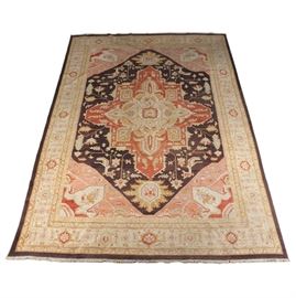 Hand-Knotted Persian Area Rug: A hand-knotted Persian area rug. The rug features a central medallion in a palette of cream, deep red, yellow and taupe that sits on top of a deep brown field. The field is covered in floral palmettes and medallions in a complementary palette with a spandrel to each corner. The field is surrounded by a compound resolved border that features a turtle pattern throughout in red, cream, and sage that sits on top of an off white field with a wool fringe to either end. The rug is not tagged.