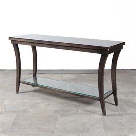 Ebonized Console Table With Glass Protective Panels: A console table in a faux walnut veneer. This table features an oversized top over block apron sides, resting on tapering saber block legs with a stretcher shelf to the bottom for support and additional storage. This table includes a full glass panel protective top and two smaller glass panels for the lower shelf.