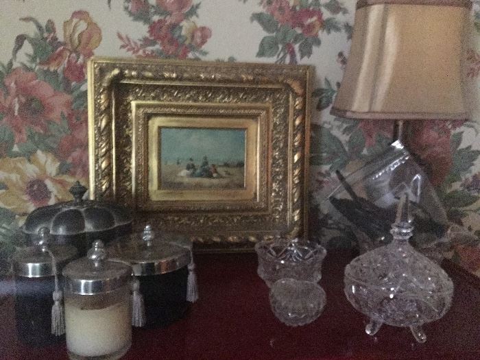 Lidded jar candles and crystal ladie's vanity ware surround a gilt-framed oil outdoor scene