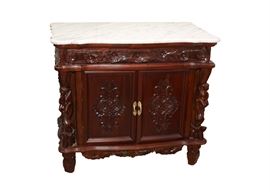 Vintage Style Marble Top Buffet: A vintage style marble top buffet. This mahogany finished buffet features a rectangular scalloped white and gray swirled marble top with an ornately carved floral top frame, two front doors and sides that feature recessed paneling and relief floral engravings that open to a shelved interior. The front sides of the cabinet feature carved figurines and front apron.