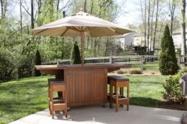 Wooden Outdoor Bar: A wooden outdoor bar. This bar features a rectangular top with drop sides,two paneled doors that open to store four stools and sits on four casters to the bottom for easy movement.Also included are six faux leather top wooden box stools and a beige canvas style umbrella.