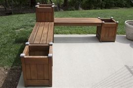 Wooden Planter Bench: A wooden planter bench. This bench features two plank style benches that are connected by three wooden box planters. Each planter box has four metal covers to the corners.