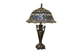 Tiffany-Style Table Lamp: A Tiffany-style table lamp. The lamp features a round, ornate footed base and is topped with a round, domed shade, made of colored leaded glass. The glass colors include pink, pink, green, blue, ruby red and lavender. The lamp has two sockets, and switches on via draw strings. The lamp is topped with a bronze-tone metal finial.
