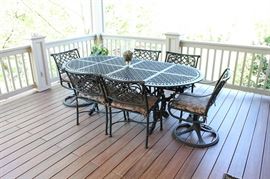 Cast Classics Outdoor Patio Dining Set: A Cast Classics, “Landgrave” collection, patio dining set with table and six chairs. There are four center chairs with four feet and arms, and two swiveling chairs with arms. The chairs feature a swirling diamond pattern to their backs and the table is oval-shaped, with an open-work, geometric pattern to its surface, and with ornately, scrolled legs and feet. Included are seat cushions with ties featuring a neutral tropical leaf pattern.