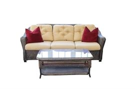 Lloyd Flanders Patio Sofa and Coffee Table: A Lloyd Flanders patio sofa and coffee table. This set is comprised of brown wicker. The sofa contains pastel yellow cushions and red accent pillows. The matching rectangular coffee table features a glass top surface and shelving below for additional storage.