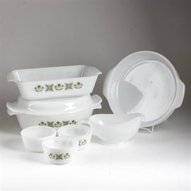 White Glass Fire-King Bakeware: A collection of white glass Fire-King bakeware. Included in this collection are a gravy boat with a handle and pour spout and a deep dish pie pan. Both of these items are plain white without a design. Also included are a bread loaf pan, an oval handled casserole and lid, and three ramekins. All of these items have a green retro design on their sides. Each piece of bakeware is marked “Anchor Hocking, Fire-King”.