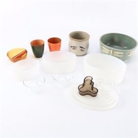 Pottery and Glass Bowls: A collection of pottery and glass bowls including a glass mortar and pestle. There is a ceramic bowl, a planter, a pair of matching bowls in asymmetrical shapes in dark and clear glass, clear glass vessels, three frosted glass bowls in different sizes, and four plastic pieces in different sizes. Many of the items are clean and unmarked, while the blue bowl has an image of a dragonfly on it in a textured ceramic material.