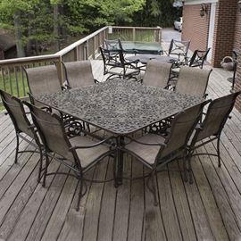 Outdoor Patio Set by Castelle: An outdoor patio set by Castelle. The set includes a large square table with a cast-iron look acanthus motif top, eight armchairs with scrolled metal frames and brown woven vinyl seats, and an umbrella base. Companion 17BAL049-040