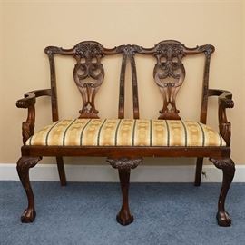 Vintage Chippendale Style Mahogany Settee: A vintage Chippendale style upholstered settee. This settee features a dark mahogany stained frame with intricate carved detailing in the splat, cabriole front legs terminating on claw and ball feet to the front. The seat is upholstered in a striped beige upholstery with leaf detail throughout. There are no visible maker’s marks or labels on this piece.