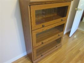 Oak three door book shelf unit; doors have inlaid glass panels for viewing, and two nobs to lift the door and roll it into the unit.