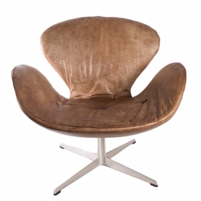Mid Century Modern Fritz Hansen Swan Chair Designed by Arne Jacobsen: A Fritz Hansen swan chair designed by Arne Jacobsen. This chair has a modern shape comprised of a rounded back with raised sides. The chair is covered in distressed brown leather upholstery with covered button accents to the seat. The chair rests on a chrome frame with a central pedestal attached to four splayed legs with capped feet. A label indicating the maker is present to the base of the chair.
