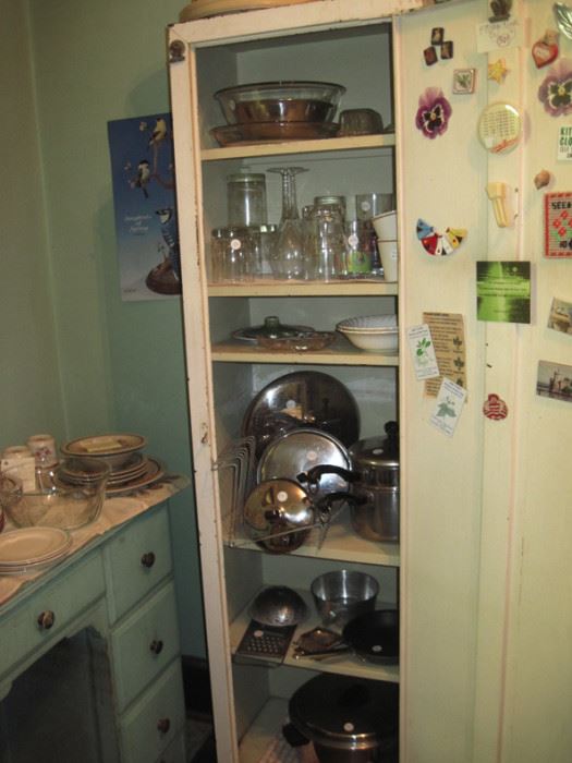 Old metal kitchen cabinet with a variety of kitchen items. Old painted wood desk with dinnerware on top.