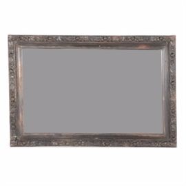 Wooden Framed Wall Mirror: A wooden framed wall mirror. The frame of the mirror is rectangular in shape with carved floral patterns throughout. The piece is dark in color with no markings.