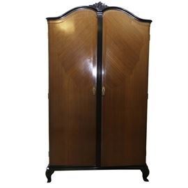 Art Deco Wardrobe: A large art deco era wardrobe. The wardrobe is constructed of wood with tiger oak veneer to the doors. The piece is trimmed in black painted accents. A floral bouquet pediment accents the top of the piece and the pieces rests on squat cabriole legs. Brass keyhole escutcheons adorn the front doors of the piece. Once opened, the piece reveals a clothing rod and bottom drawer accented with brass pulls.