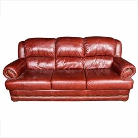Contemporary Brown Leather Sofa: A contemporary brown leather sofa. The sofa features three attached, plush pillow backs, with wide, rolled arms, and three contoured box seat cushions. The fronts of the arms and the front of the base are decorated with brass tacks, and the sofa rests on round, short wood bun feet. The sofa is fully upholstered in cognac colored leather, and is not marked.