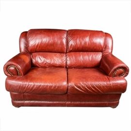 Contemporary Leather Loveseat: A contemporary brown leather sofa. The love seat features two attached, plush pillow backs, with wide, rolled arms, and two contoured box seat cushions. The fronts of the arms and the front of the base are decorated with brass tacks, and the sofa rests on round, short wood bun feet. The sofa is fully upholstered in cognac colored leather, and is not marked. This item matches item 17DEN038-006.