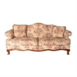 Chippendale Style Camelback Sofa by Schnadig: A Chippendale-style camelback sofa by Schnadig. This sofa features a serpentine back leading to scrolled arms surrounding a pair of oversized seat cushions over an upholstered seat rail over a scalloped apron, resting on cabriole legs with carved knees ending in claw and ball feet. The sofa includes a cream hue field with a floral stripe print and piping detail accents. There are coordinating throw pillows included and a tag which reads “Schnadig” to the interior of the seat decking. For coordinating items, please see 17BOS067-103 and 17BOS067-107.