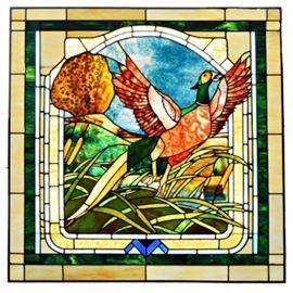 Stained Glass Window with Pheasant: A large stained glass window featuring a startled pheasant flying out of a a bed of cattails at the edge of a pond with bright blue sky in the distance. The image is surrounded by a white with blue and green arched frame of stained glass.