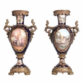 Pair of Porcelain Urns with Figural Handles: A pair of rather large reproduction mounted porcelain urns featuring very ornate cast metal mounts, figural handles and footed bases all with a brass plated finish. The cobalt blue porcelain body features a main transferred image of a Provincial social gathering and cottage landscape scene to the verso with smaller vignettes of country cottages to the neck and base. The urns are embellished with hand painted gold accents. Unmarked.