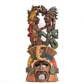 Carved Mayan Warrior Mask: A Mayan wooden warrior mask. Carved from tropical wood, this piece features a victorious warrior in full battle regalia of a feathered head dress, beads and cloth, kneeling with the head of his vanquished foe held up for inspection. A panther figure appears to be coming from the top of the victim’s head, while an opened mouth snake slithers beneath. The figures are resting on a series of rectangular pieces lined in gold with a central, crescent moon figure with human face. The mask ends with an opened mouth Mayan warrior face with flanking adornments. The mask hangs with a wire to verso. This item matches 17COL081-086 in this sale.