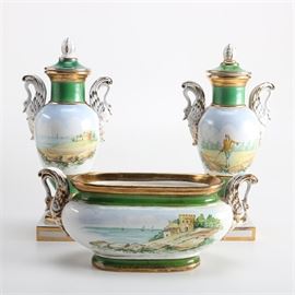 Garniture Set: A garniture set. This set includes two urn style pieces and a jardiniere. Each piece is detailed with a green and gold tone border, centering a colored landscape scene, and gold tone accented side handles and pedestal base.