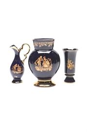 Cobalt Blue and Gold Tone Porcelain Vessels Featuring Limoges Pieces: A group of blue and gold tone décor pieces featuring Limoges pieces. This includes an oil style pitcher and round vase with matching Colonial-era motifs with gilding. There is also a footed vase with floral transfer prints and gilding with an indiscernible marking to the underside.
