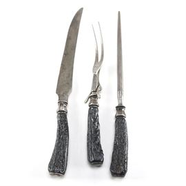 Steel Carving Set: A three piece carving set. Featuring stainless steel with carved wood handles resembling bone, this set features a knife, a sharpener, and a carving fork. The knife features an illegible mark at the base of the blade.