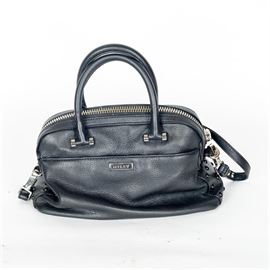 Milly Black Leather Handbag: A Milly black leather handbag. This handbag has a black leather exterior with hand and shoulder straps. It has a top zipper closure with and exterior back pocket, and an interior zipper pocket.