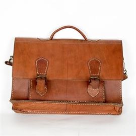 Vintage Leather Briefcase: A vintage leather briefcase. This briefcase has a brown leather exterior with a shoulder strap and handle. There are two brass buckle closures, a zippered front pocket, and an open front pocket. There are two large open interior sections.