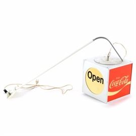 Hanging Coca-Cola Open Sign: A hanging Coca-Cola lighted “Open” sign. This cube sign has a metal arm that attaches to the wall, through which the cord runs into the metal cube housing. Two circular cut-outs read, “OPEN” with the slogan, “Enjoy Coca-Cola” on the adjacent sides. A switch on the cord allows for turning the light on and off.