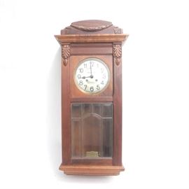 Besanger Selecta Wall Clock: A Besanger Selecta wall clock. The clock is cherry wood that features a rectangular design and is topped with a carved stand up bonnet and carved corner blocks that flank a brass bezel on a white dial with black Arabic numbers. Other features include raised glass panels that sit on a plinth style wood shelf and tapered base. The clock is marked “Besanger Selecta”.