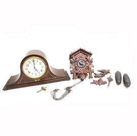 Weight Driven Cuckoo and Mantel Clock: A Plymouth mantel and German cuckoo. The selection features a mantel clock that is wood and features a curved shape on a plinth style base with a gilded design and bezel over a white face with black Arabic numbers and hands. The cuckoo clock features a wood housing with carved wood accents depicting birds and leaves, having a carved leaf pendulum. The cuckoo bird is plastic and the mechanism is weight driven by two cast metal pine cone weights.