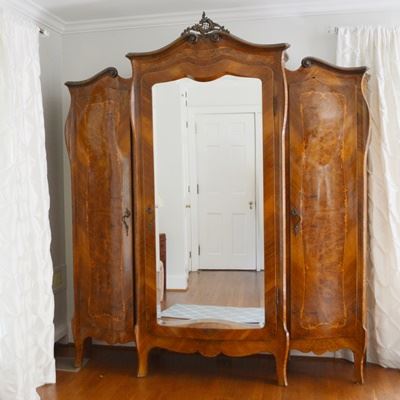 Antique Italian Louis XV Style Serpentine Wardrobe: An antique Italian Louis XV style serpentine wardrobe cabinet. Designed as a three door armoire, this set features a curved scroll crown, marquetry inlay, burled walnut veneers, and other veneers on the curving panels. The center unit stands proud of the side cabinets, includes a beveled door mirror, with a bronze detail above and ornate pull hardware. All cabinets feature a distinctive blade style flared foot and scalloped aprons. The poplar interiors feature an inlay border on all panels. The left unit includes two shelves, the center unit a wardrobe bar and lower drawer with dovetail construction, and the right unit a wardrobe bar. A working key is included. This unit breaks into five pieces with metal pins for removal. This unit coordinates with #17CIN294-039 dresser, #17CIN294-032 end tables, #17CIN294-036 beds, and #17CIN294-033 vanity.