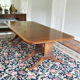 Vintage Duncan Phyfe Style Dining Room Table: A vintage Duncan Phyfe style mahogany table. The mahogany table features rounded ends with a wood veneer banded edge. The table includes two additional leaves, but it is shown at its full length. Supported by two reeded panels, the double reeded and splayed legs with brass feet are connected with a turned spindle. An additional panel and leg is designed into each end to allow the table to expand and accommodate additional length. The table is thought to be Baker but is unmarked.