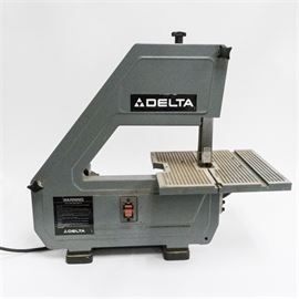 Delta Band Saw: A Delta10" band saw. The saw is metal with a gray and black finish and stands on four black feet. The saw has a metal table with tension adjustment, red power switch and locking capabilities, blade direction, and a black electrical cord. The saw reads “Delta International Machinery Corp.”.