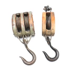 Wooden and Metal Industrial Pulleys: A pair of industrial pulleys made of wood and metal. Both pulleys, or blocks, feature wooden cases with metal hooks and hardware. One bears remnants of an old grey tone paint job.