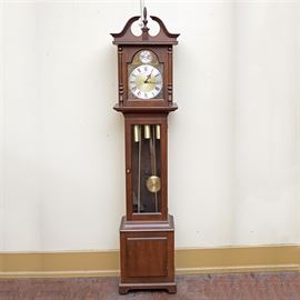 Barwick "Tempus Fugit" Grandmother Clock: A Barwick Tempus Fugit grandmother clock. This selection showcases a wooden frame with arched detailing and a finial to the top. The face is bordered by gilded metal adorned with foliates and an illustration of the world with the words “Tempus Fugit” displayed in a figural ribbon. It also features a glass door showcasing the pendulum and is set upon a wooden base. It is labeled by Barwick.