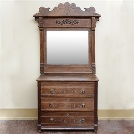 Victorian Eastlake Style Dresser With Mirror: An antique Victorian Eastlake style dresser with mirror. The mirror is presented in a walnut frame, matching the dresser. Its crest is serpentine with lightly incised floral and scrolling lines above an applied floral carving. The carving is flanked by wheat carvings and scrolled supports. The walnut dresser features three dovetailed drawers with lightly incised floral and scrolling lines. Each drawer has two ring pulls with silver tone geometric backplates and a matching center escutcheon. It sits on caster feet.