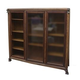 Vintage Bookcase With Lion Head Accents: A vintage bookcase. This rectangular top piece features three cabinet doors with clear glass panels opening to adjustable shelves with a thin apron terminating on claw feet. This piece features a lion head to the top of each pilaster. There are no visible maker’s marks.