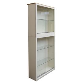 Contemporary Glass Fronted Stackable Cabinets: A pair of stackable glass fronted display cabinets. Both display modern frames with a cream laminate finish, and sliding glass doors on chrome tracks. The larger cabinet had two adjustable glass shelves, and the smallest and one. No discernible maker’s mark was found.