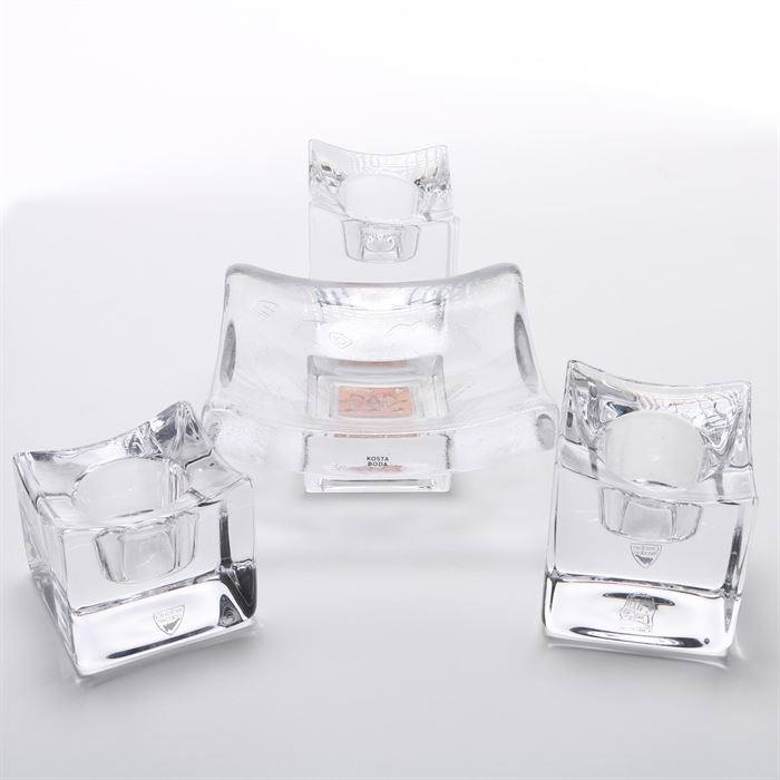 Orrefors Sweden Candle Holders and Kosta Boda "Dad" Domino Holder: A collection of three glass candle holders and a domino holder. This assortment features three square translucent glass candle holders labeled “Orrefors Sweden” and a square art glass domino holder by Kosta Boda marked “Dad” to the center. The candle holders are designed for single candlesticks, and vary in size.