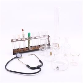 Chemistry Set: A chemistry set with assorted beakers. This set features a selection of clear glass test tubes with a metal holding rack and beakers of various size and shape including two hand blown Pilgrim Glass beakers. Also included is a stethoscope.