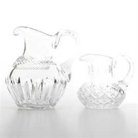 Crystal Water Pitchers: A pair of crystal water pitchers. The pitchers in this lot are made of clear glass, one with a cut, fluted design along with a wraparound fern pattern, the other featuring a diamond and palmette pattern. The items are unlabeled.
