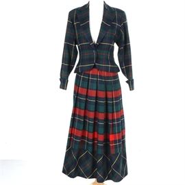 Circa 1980s Perry Ellis Plaid Wool Skirt Suit: A circa 1980s plaid wool skirt suit by Perry Ellis. This suit includes a blazer and a midi skirt composed of plaid worsted wool in shades of navy, red, green, and yellow. The blazer has a notched lapel, buttoned cuffs, and a single button closure. The skirt is pleated. Both pieces are tagged “Perry Ellis”.
